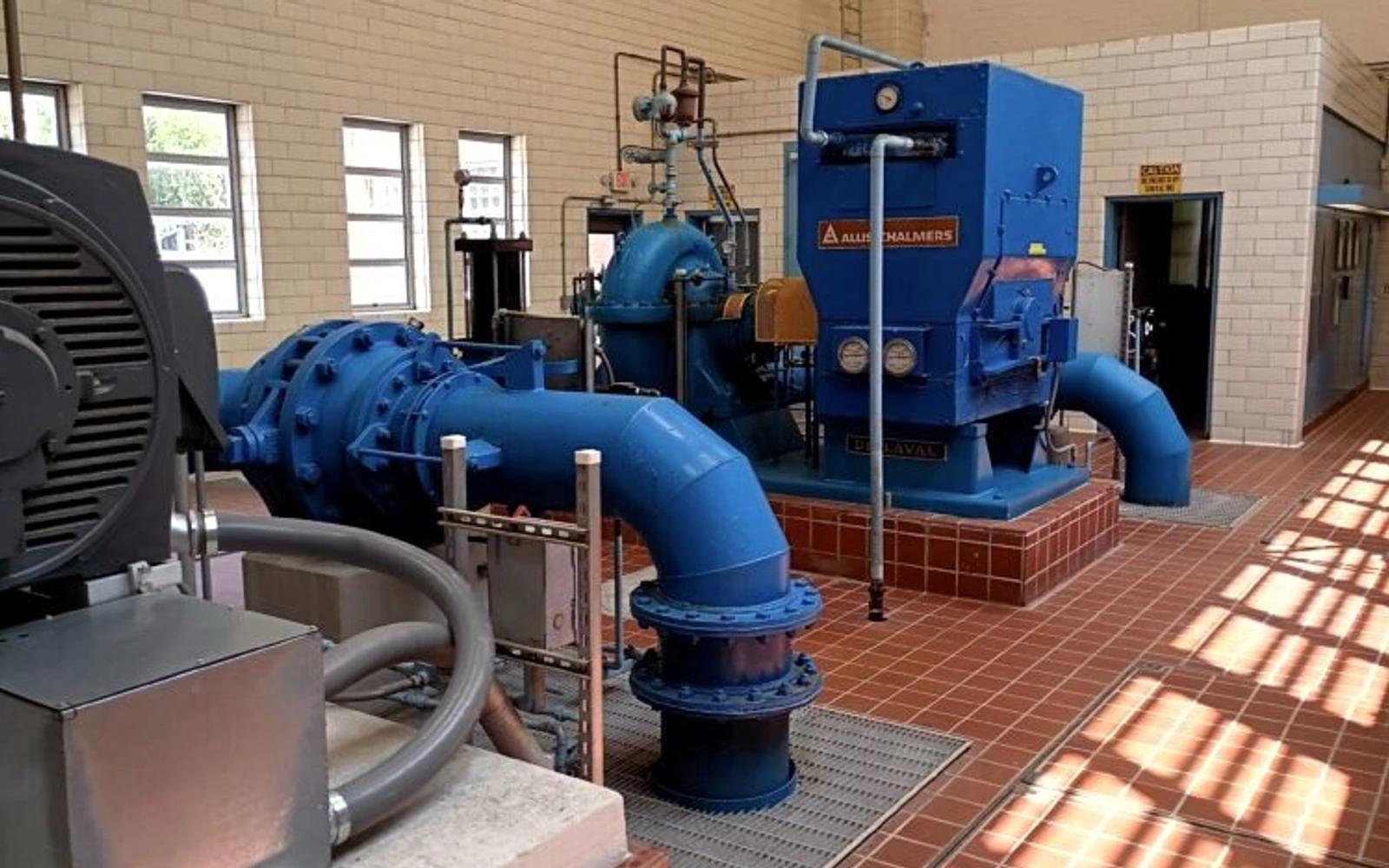 Rows of blue industrial pumps and pipes fill the tiled interior of Adamsville Pumping Station; sunlight falls through high windows onto the floor.