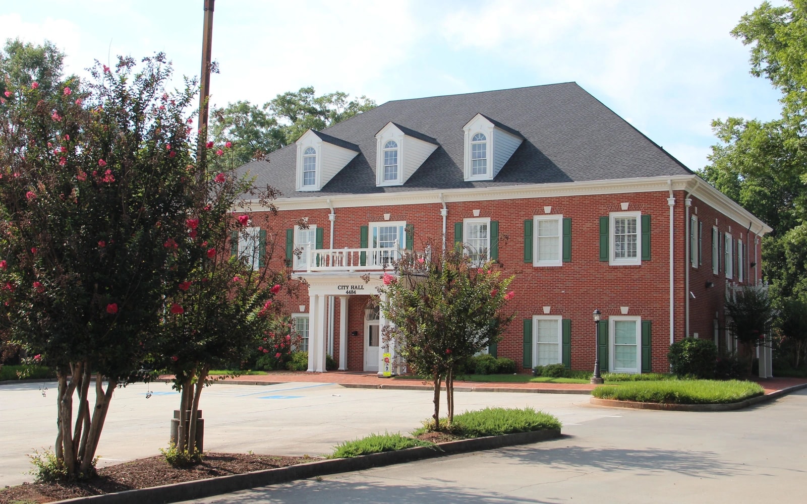 Red-brick Powder Springs City Hall with white columns, dormer windows and trees beside an empty paved parking area.
