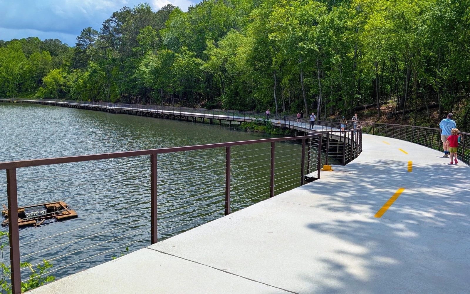Walkers follow a concrete boardwalk curving along Orkin Lake, with metal railings beside the water and a wooded shoreline beyond.