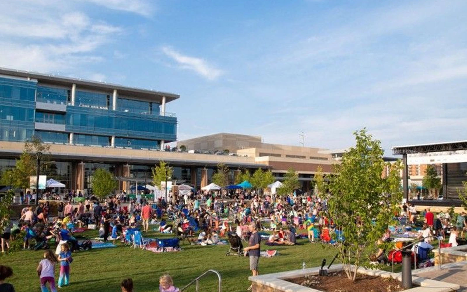 A crowd gathers on the lawn at City Green, with City Springs buildings and an outdoor stage behind them.