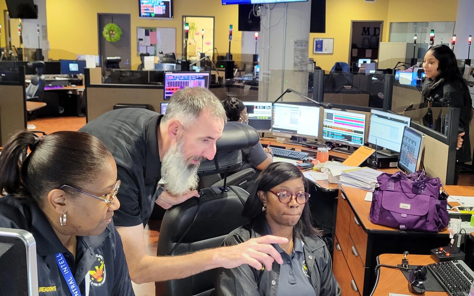 Three Atlanta Police Department staff members confer beside dispatch consoles and multiple monitors in the E-911 communications center; other workstations are visible behind them.