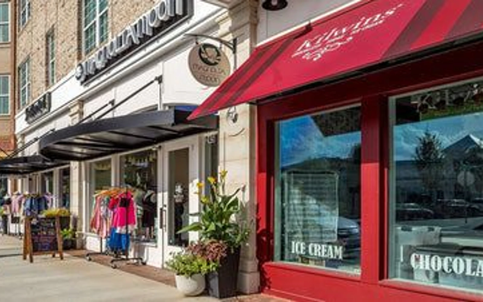 Boutique and neighboring shop fronts along a downtown Alpharetta sidewalk, with clothing racks outside and a red-awning storefront in the foreground.