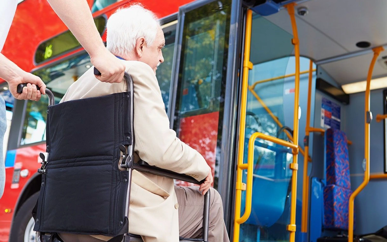 An older adult in a wheelchair is assisted toward the open doorway of a red-and-blue city bus; the image is representative, not a Fulton County service vehicle.
