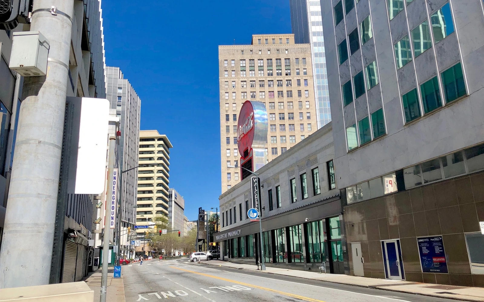 A downtown street lined with multistory buildings, with lane markings leading toward the distance beneath a blue sky.