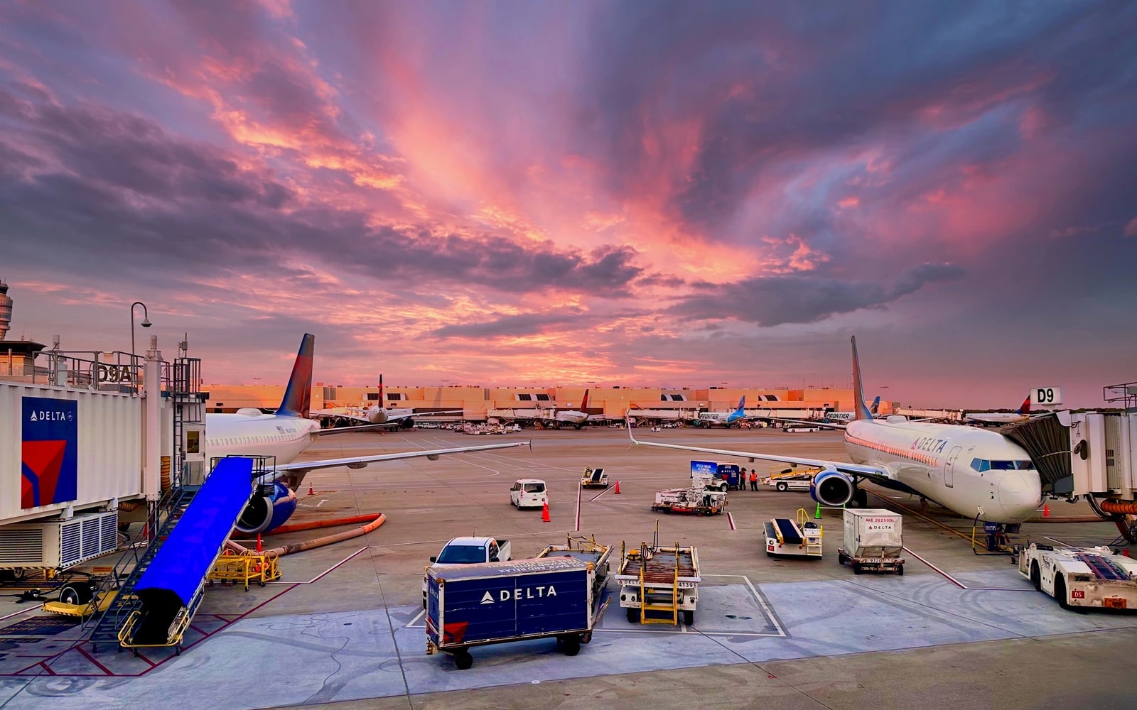 Passenger aircraft beside boarding bridges, with baggage carts and service equipment on the airport apron beneath a pink and purple sky.