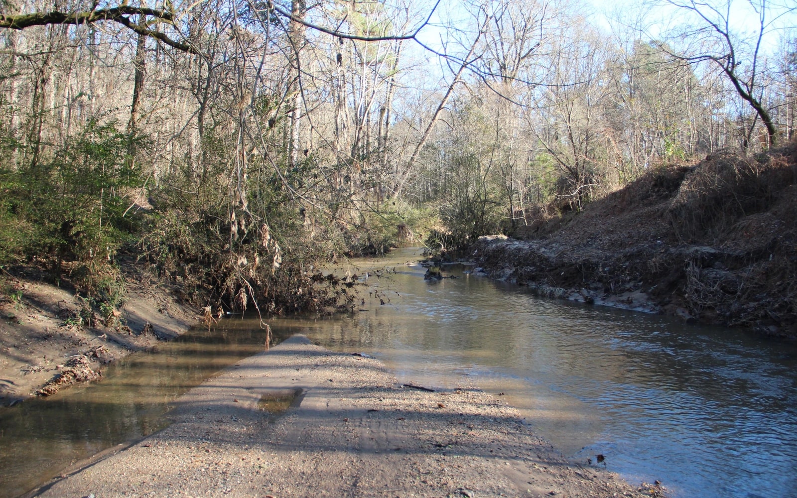 A shallow creek curves between wooded banks, with a gravel bar in the foreground and mostly leafless trees overhead.