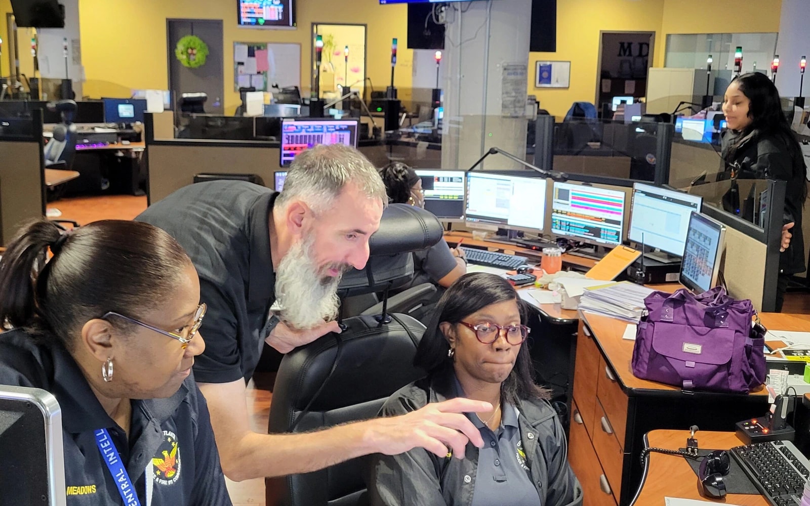 Several E911 center staff gather around computer dispatch screens; a bearded man points at a monitor while two uniformed women look on, with more workstations behind them.