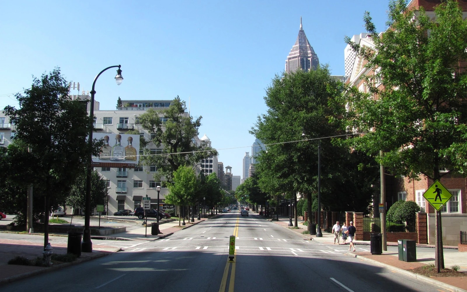 A broad paved street with lane markings runs between tree-lined sidewalks and multistory buildings, with a pointed tower rising in the distance.
