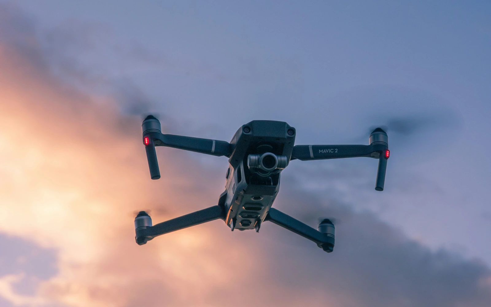 A camera-equipped quadcopter seen from below against pink and blue sunset clouds.
