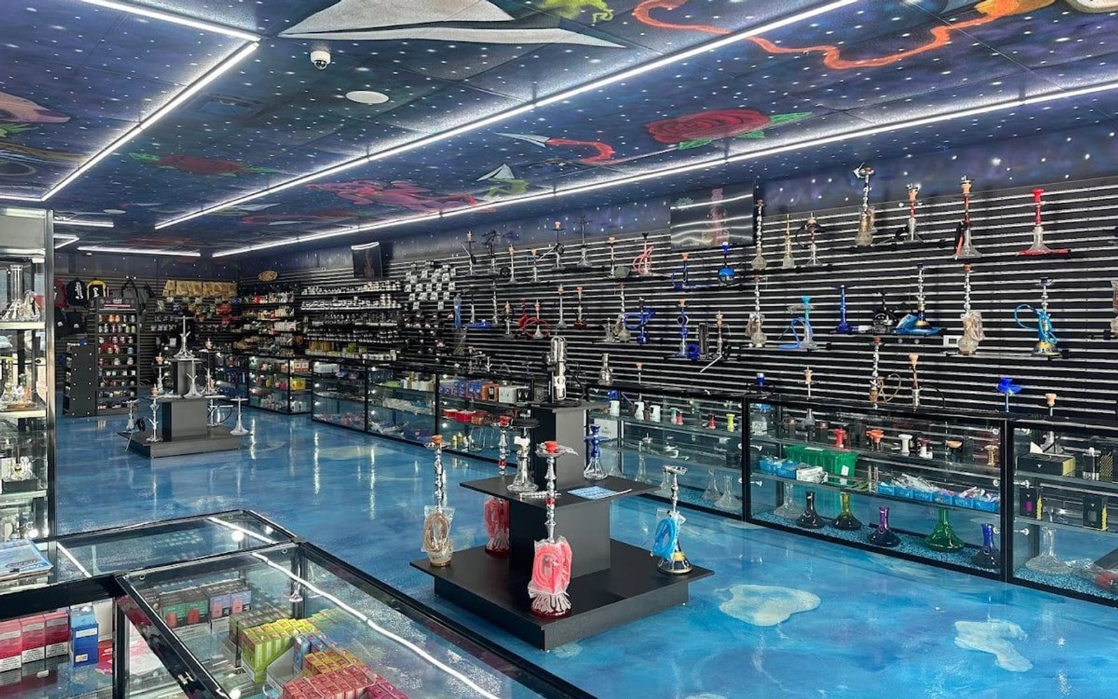 Rows of glass pipes and water pipes fill illuminated display cases beneath a colorful, space-themed ceiling inside a Sandy Springs smoke shop.