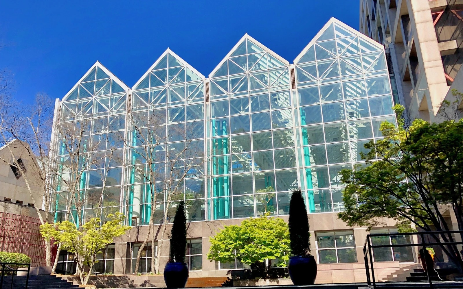 The Fulton County Government Center's glass atrium facade rises between concrete office wings beneath a clear blue sky, with trees and planters along the plaza.