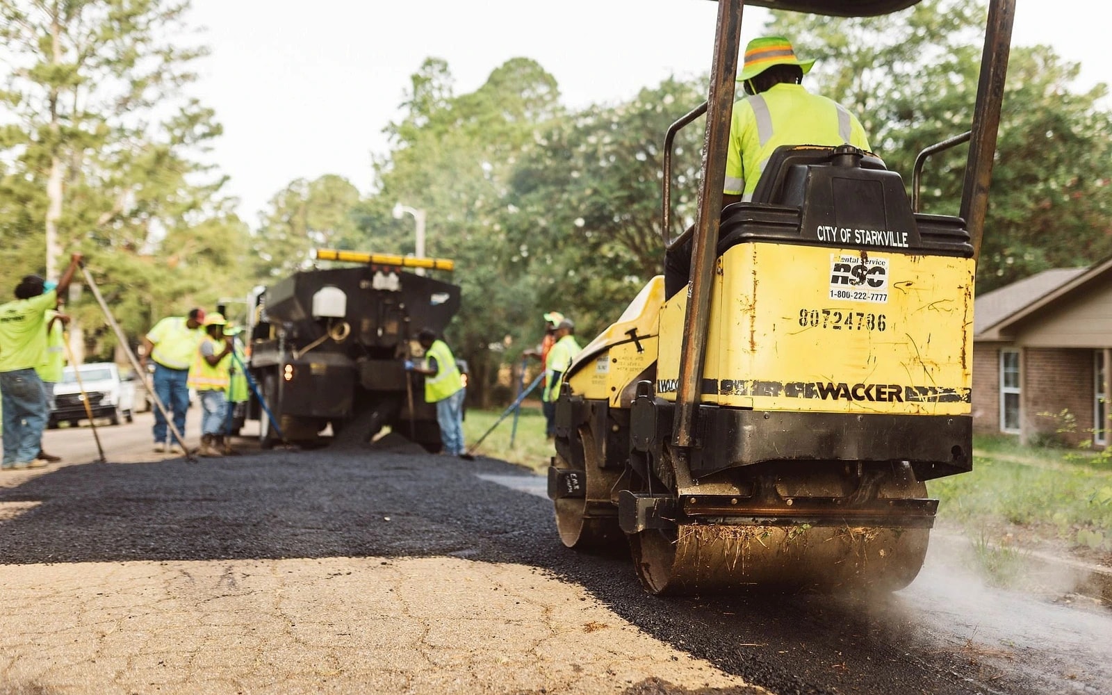 A city worker operates a road roller in the foreground as a crew lays dark pavement on a residential street.