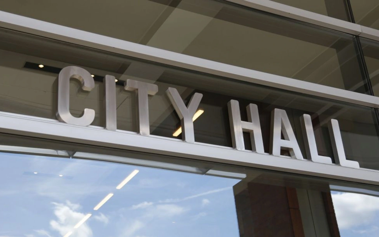Close view of raised CITY HALL lettering above a glass entrance, reflecting clouds and lights.