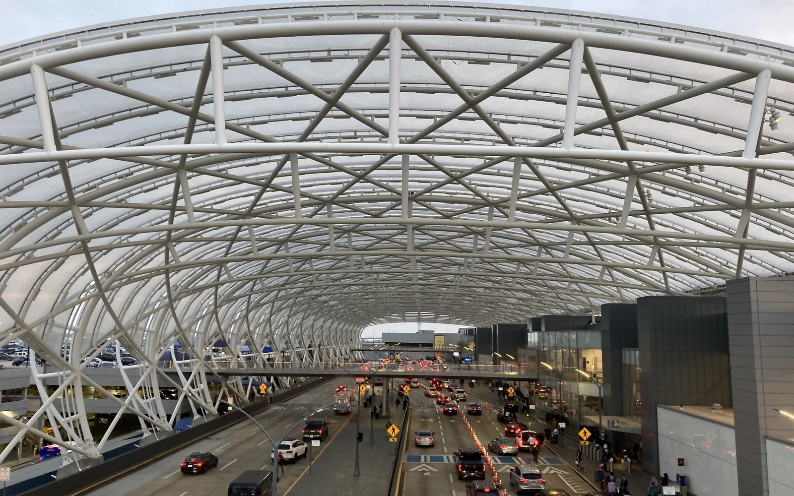 Cars line airport terminal pickup and drop-off lanes beneath a broad, curved canopy supported by white steel trusses.