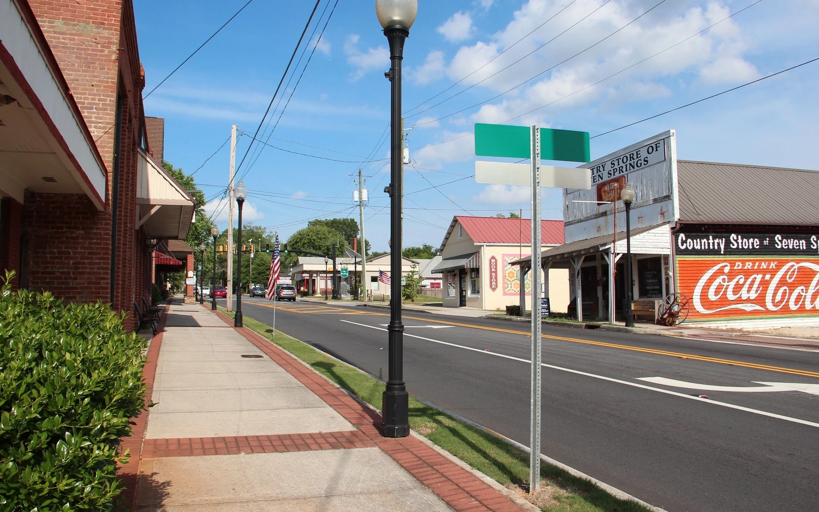 A sidewalk edged with brick and shrubs runs beside a downtown street, with a decorative lamppost, overhead utility lines and storefronts across the road.
