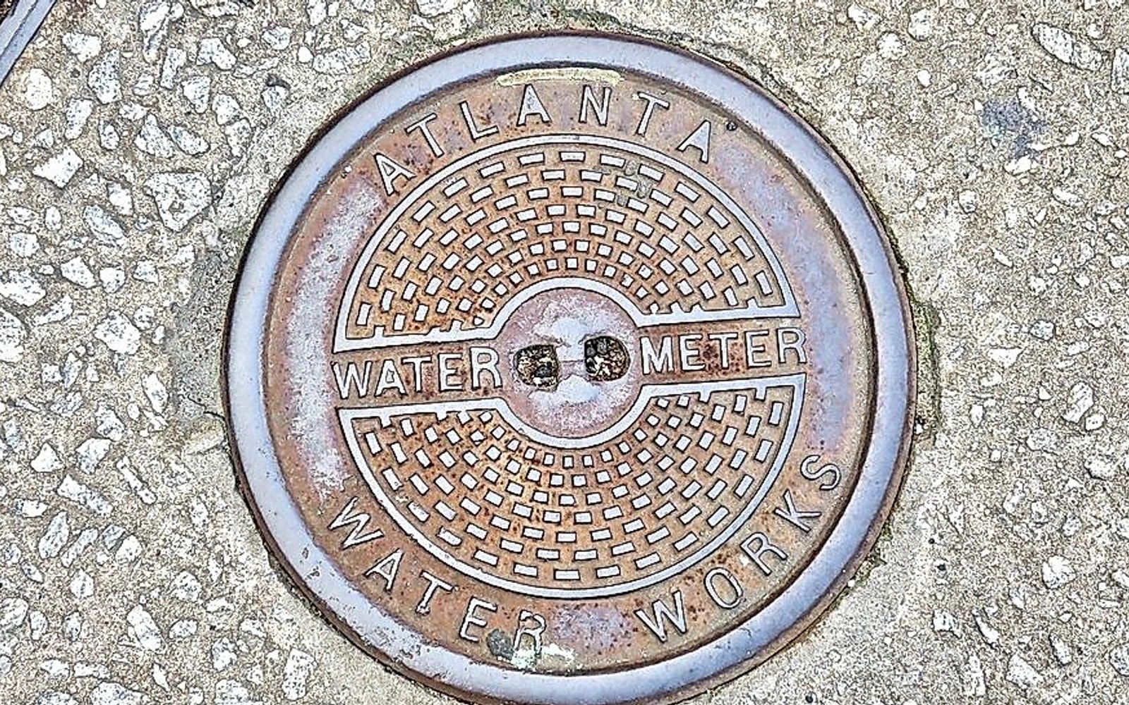 A round metal Atlanta Water Works water meter cover is set into a rough concrete sidewalk.